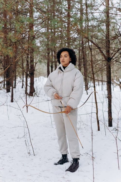 a young woman with curly dark hair stands in a snowy forest, holding a wooden bow and arrow. she is wearing a light beige puffer jacket and matching pants, along with dark boots. the snow covers the ground, and the trees are bare, indicating a winter scene. the woman appears focused, as if preparing to shoot an arrow. the overall atmosphere is serene and peaceful.