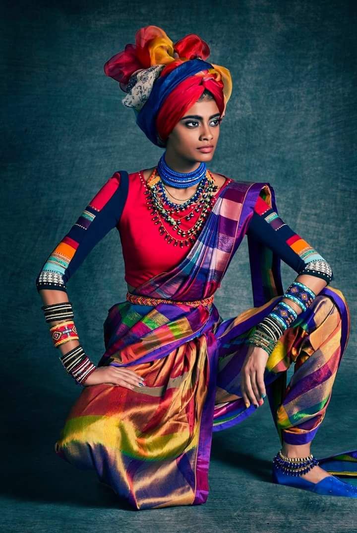 a woman in a colorful saree and jewelry is posed in a studio setting with a colorful turban. she is looking to the left with a serious expression.