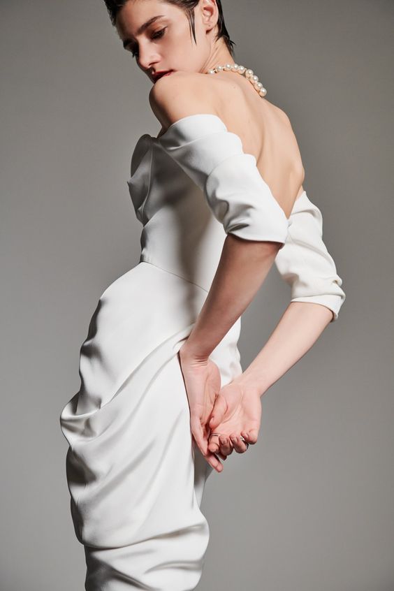 a woman wearing a white offtheshoulder draped dress stands against a light gray background, gazing down at the skirt. she wears a pearl necklace and her hair is styled in a sleek, wet look. the lighting is soft and natural, creating a clean and minimalist aesthetic.