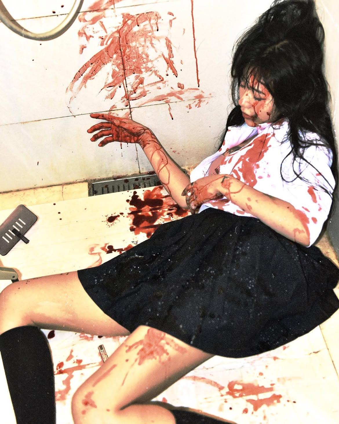 a woman with dark hair, wearing a school uniform, is lying on the floor in a bathroom, covered in blood. there is blood splattered on the walls and floor around her, suggesting she has suffered injuries. the scene appears to be a crime scene.