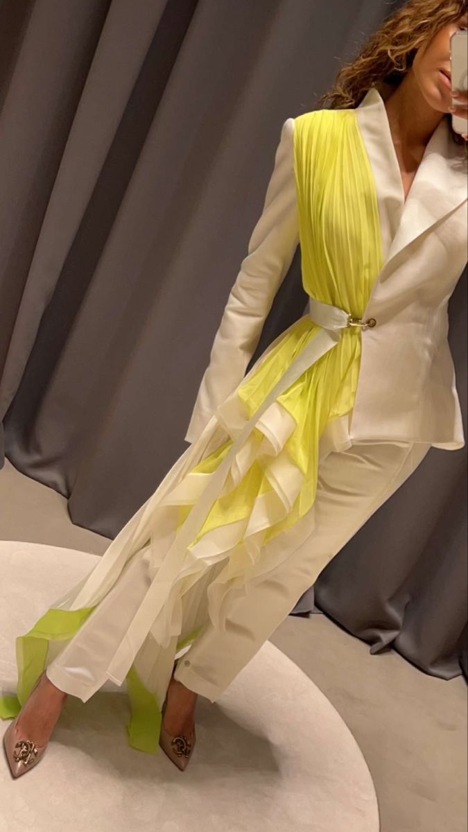 a woman is wearing a white blazer and pants with a yellow pleated sash draped over one shoulder, secured with a white belt. she is also wearing pointedtoe stilettos with chanel embellishments. she is in what appears to be a fitting room, standing on a circular platform.