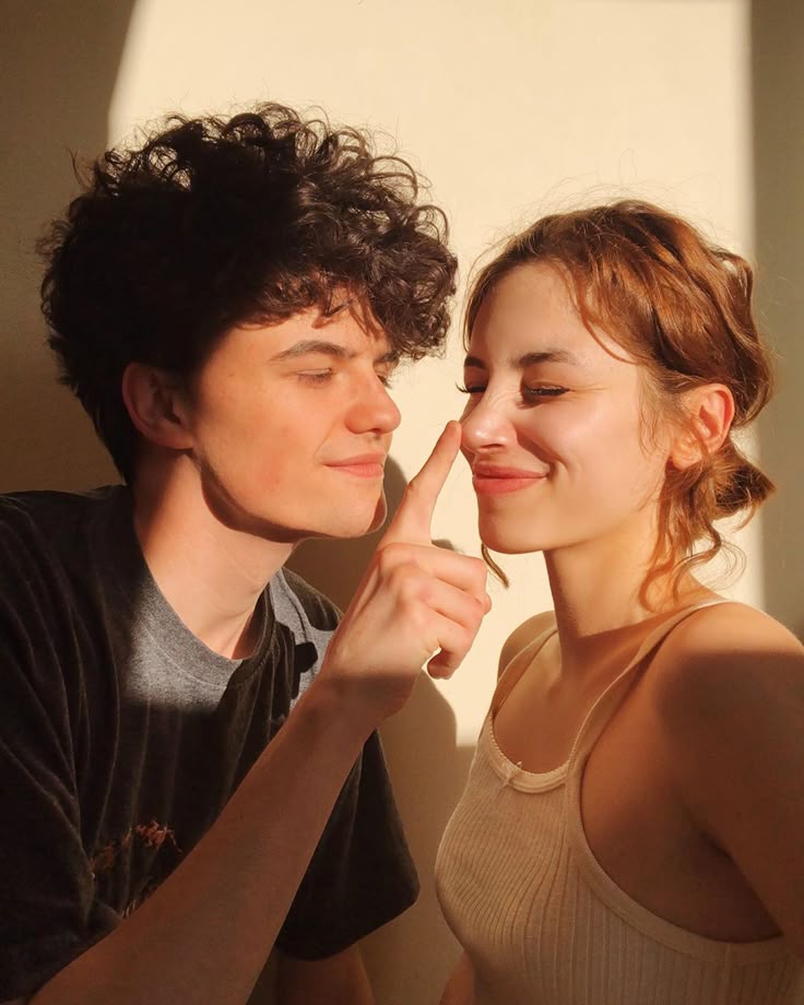 a young couple is posing indoors in warm sunlight. the man has dark curly hair and is wearing a gray tshirt. he is gently touching the woman's nose with his index finger. the woman has reddishbrown hair pulled back and is wearing a beige tank top. they are both smiling slightly, and the scene conveys a sense of affection and closeness.