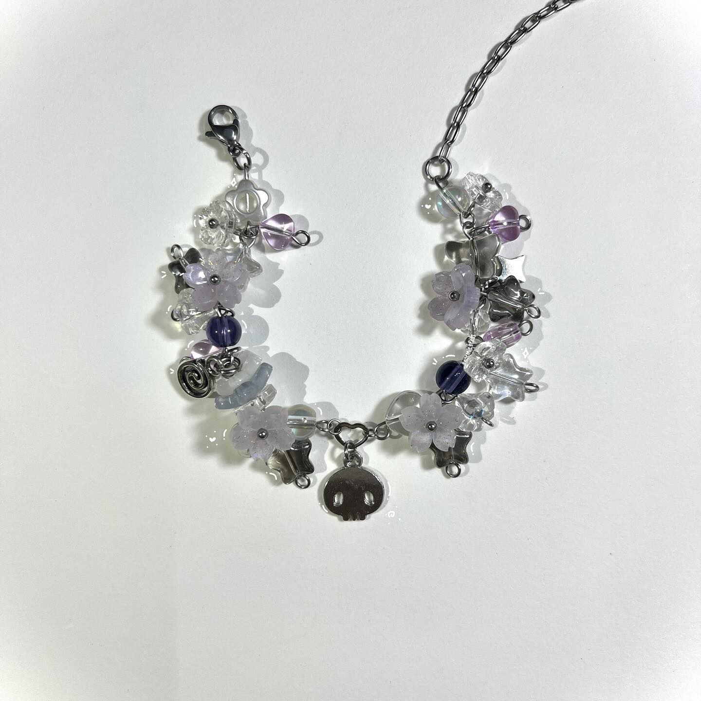 a closeup shot of a beaded bracelet with various charms and a silver chain. the bracelet is adorned with flowershaped beads in white and light purple, interspersed with clear and dark purple round beads. it features charms such as a skull, silver stars, and other decorative elements. the bracelet is presented on a white surface with soft lighting. the clasp is visible on one end, and the chain extends upwards from the bracelet.