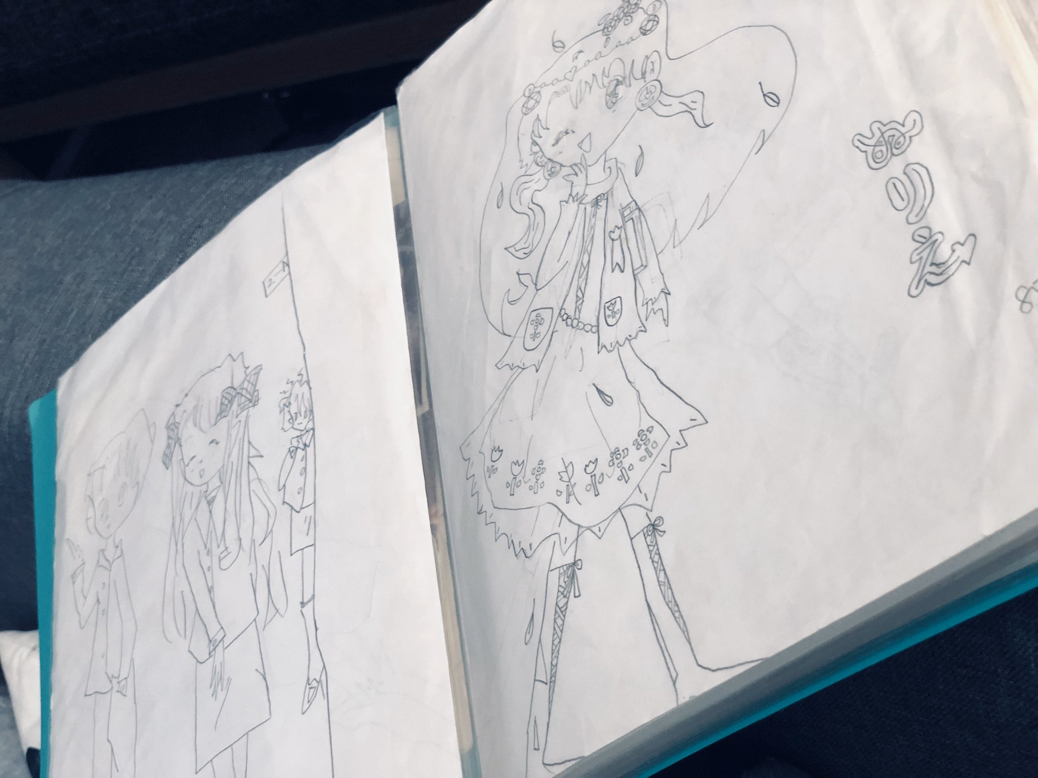 the image shows two pages from a sketchbook open to show pencil sketches of a female anime character. on the left page, there are two smaller sketches of the same character. on the right page, there's a larger, more detailed sketch of the same character in a dynamic pose. the drawings are in black and white and depict a girl wearing a dress and boots with floral decorations. she has long hair and is holding a flower in her hand. the sketchbook is teal.