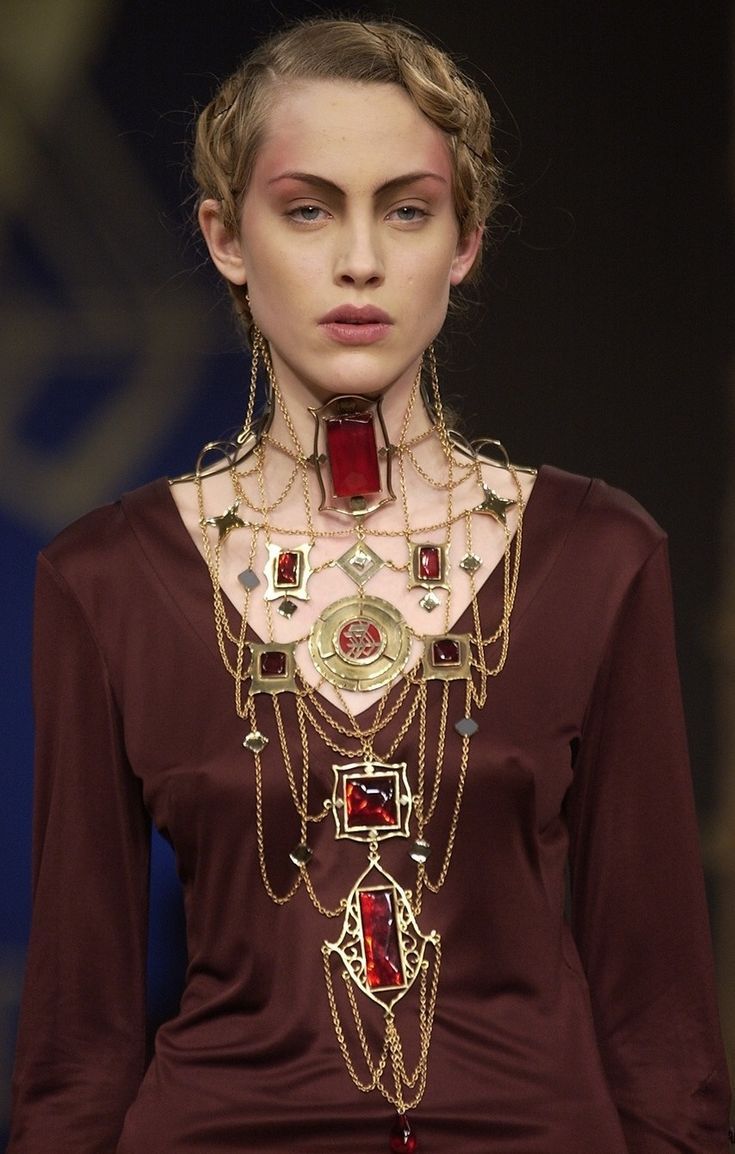 a woman with her hair up in twisted braids and striking makeup is wearing an elaborate gold necklace with red gems, which she is modeling with a matching maroon dress in a fashion show on a runway.