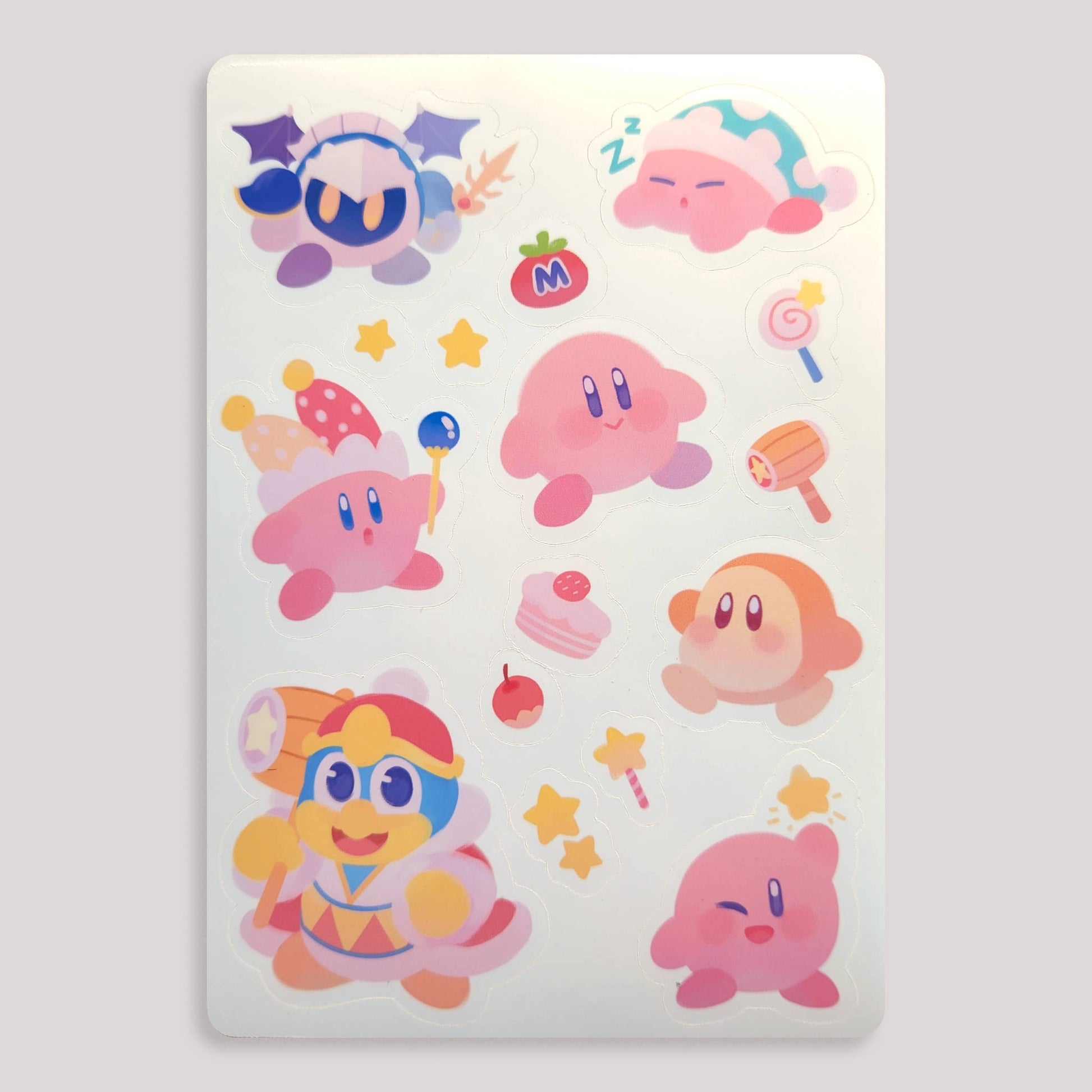 the image shows a sticker sheet featuring various cute, pastelcolored illustrations from the kirby video game franchise.  the sheet includes kirby in several different forms (sleeping, wearing a jester hat, etc.), meta knight, king dedede, and other supporting characters.  the art style is soft and rounded, with a focus on pink and other light, pastel colors.  the stickers are arranged in a pleasing, somewhat random, but cohesive manner on the sheet.  small details like stars, sweets, and a mallet add to the overall whimsical charm of the design.  the sheet is rectangular and appears to be printed on a glossy material.