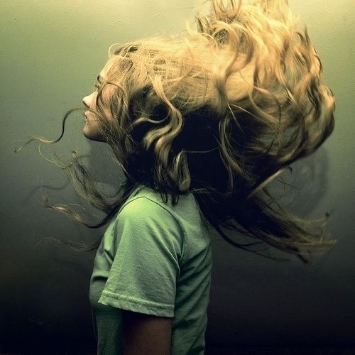 the image is a portrait of a woman with long, curly blonde hair. the woman is shown in profile, with her head tilted back and her hair flowing behind her. she is wearing a green shirt. the background of the image is a dark, muted green. the lighting in the image is soft and diffused, creating a dreamy and ethereal atmosphere. the overall effect of the image is one of movement and energy.