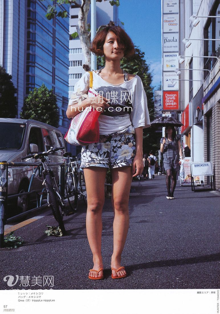 a young woman stands on a city sidewalk, posing for a street style photo. she wears a white graphic tshirt, floral shorts, and brown flipflop sandals. she carries a red tote bag over her shoulder. the background features a street scene with buildings, vehicles, and other pedestrians.