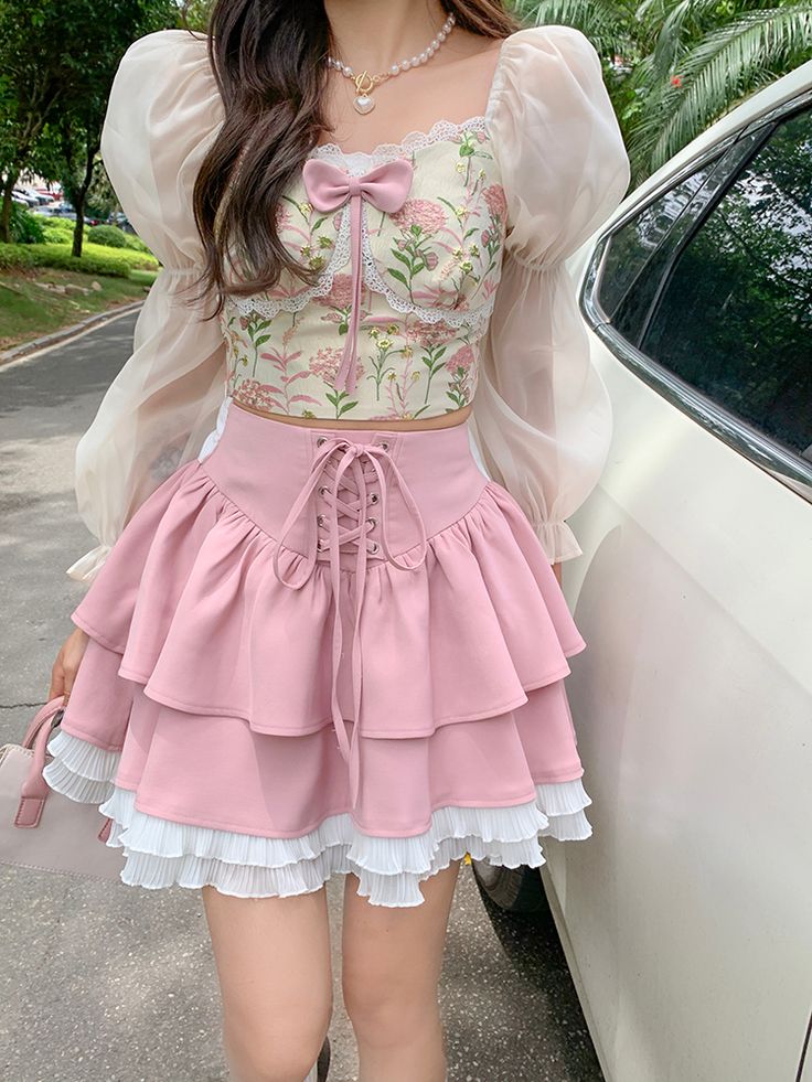 a young woman is standing in the street, wearing a pink and white lolita fashion outfit. she is wearing a pink floral crop top with puff sleeves, a pink tiered skirt with lace trim, and white shoes. she is also carrying a small white purse.  she is standing in front of a white car.