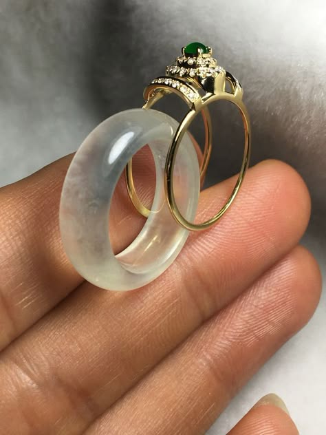 a close up of a jade ring with a gold band and a crownlike setting with a green gem stone and diamonds. the ring is set on top of a white jade ring. the rings are sitting on a person's finger. the image is well lit and in focus.