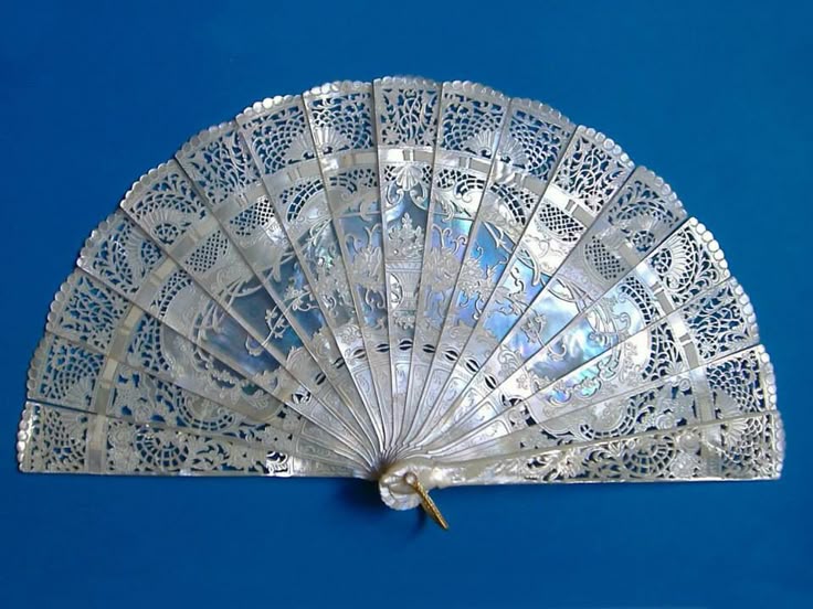 the image shows an ornate, vintage hand fan made of mother of pearl. it features intricate filigree designs and a decorative pattern. the fan is fully opened, displaying its delicate structure and iridescent sheen, and it is placed against a solid blue background.