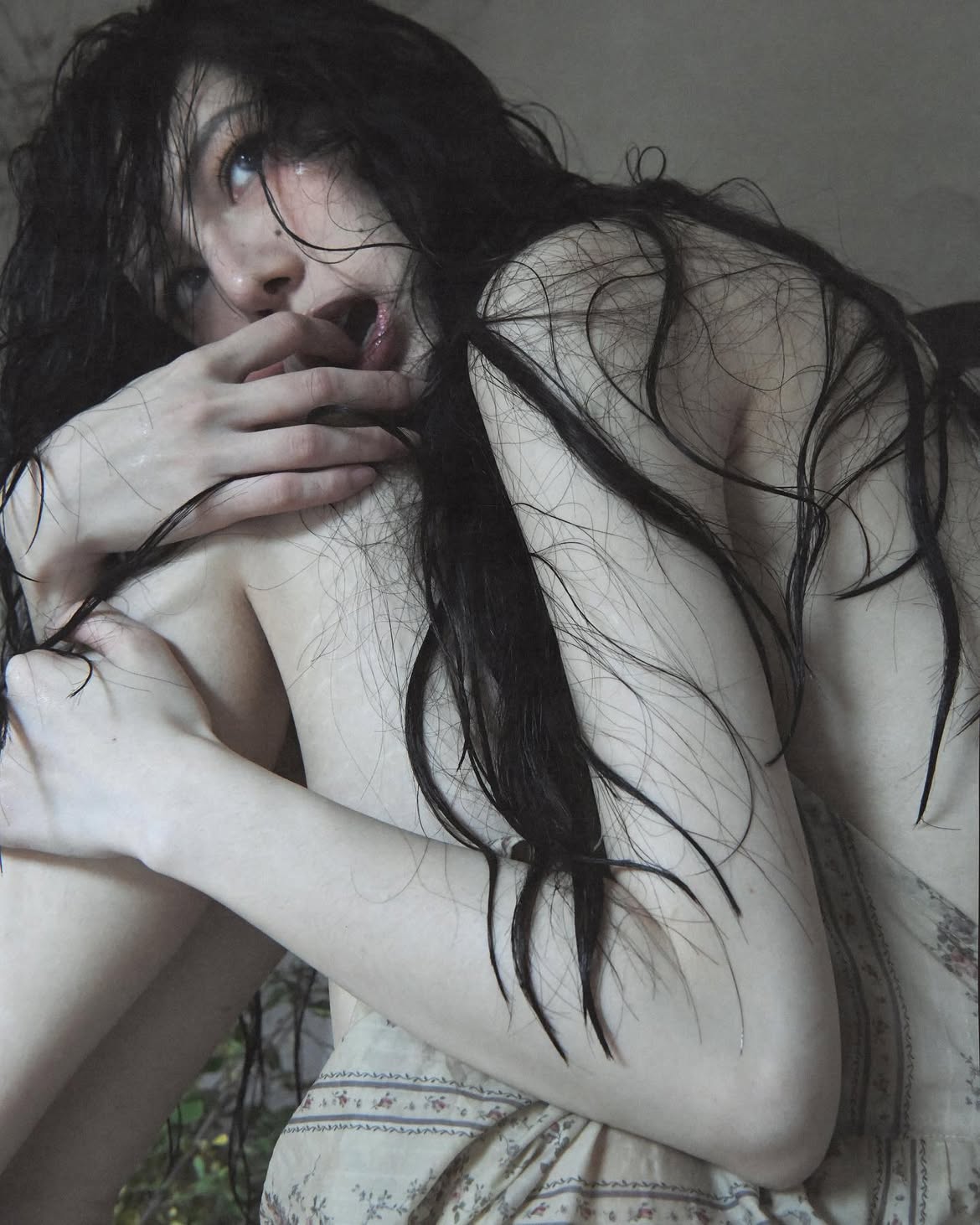 a portrait of a woman with long, black, wet hair, bare shoulders, and pale skin. she is sitting down, with her knees pulled up towards her chest, and is looking up with an open mouth. her fingers are visible and her expression is sensual and emotive. she is wearing a patterned dress. the overall mood is moody and aesthetic.