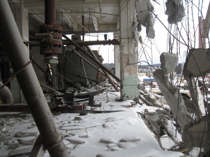 he image shows the aftermath of a building collapse.  the interior of a large industrial structure is depicted, with large sections of concrete flooring and walls destroyed and strewn across the snowcovered ground. twisted metal pipes, wires, and rebar are visible amongst the rubble. the remaining structure shows significant damage, with exposed interiors and broken supports. snow covers the debris field, indicating a winter setting. the overall scene conveys a sense of extensive destruction and abandonment.