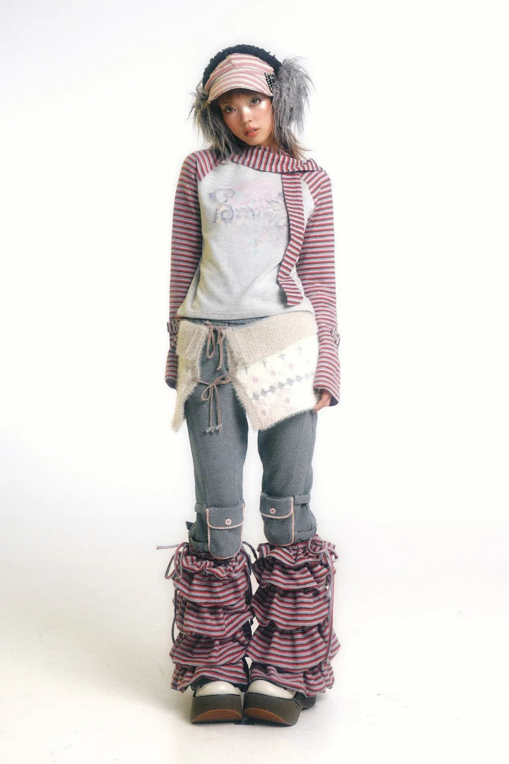 full shot of a young woman, possibly a fashion model, posing against a white background. she is wearing a unique and layered outfit that combines several different textures and patterns. she wears a pink and white striped hat with feathers, a light gray and pink striped longsleeved shirt, a  light gray and offwhite knitted wrap, grey pants with pink accents on the cuffs, and layered pink and grey striped leg warmers.  her overall style is eclectic and blends elements of casual and quirky fashion.  the image is professionally taken and likely intended for a fashion publication or online store.
