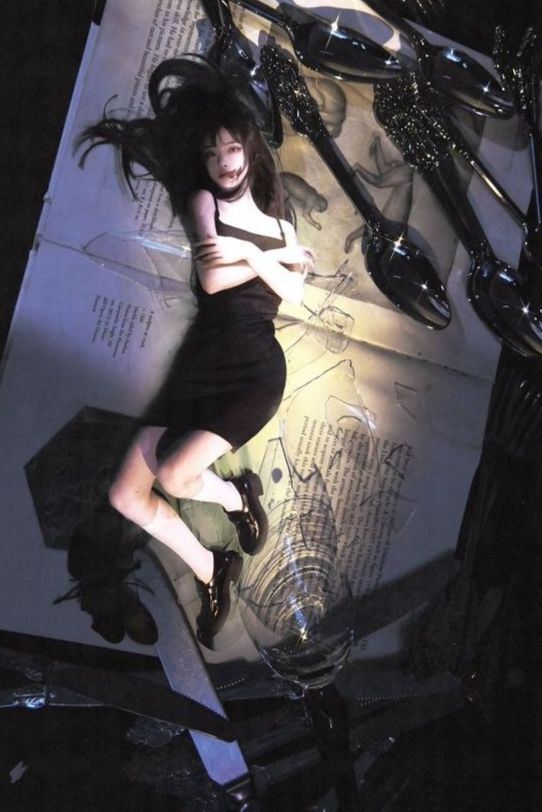 a young woman with long black hair is lying on a surface covered in fragmented book pages and shattered glass. she wears a black dress and platform shoes, her body positioned amidst the scattered debris. the scene is dramatically lit from above, casting strong shadows and creating a dark and evocative atmosphere.  the overall aesthetic leans towards grunge and alternative fashion.