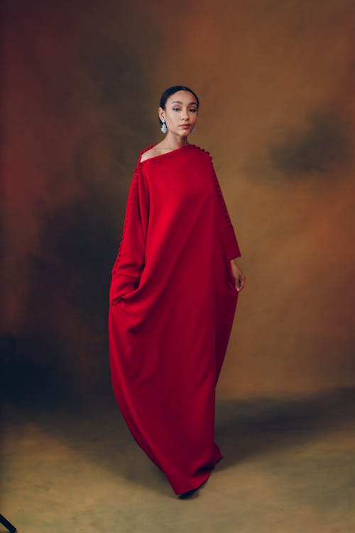 a woman in a red, oneshoulder kaftanstyle gown stands in a studio setting. she is facing the camera and is wearing large, teardropshaped earrings. the background is a mottled mix of brown and red tones. the dress is floorlength and has a loose, flowing fit.