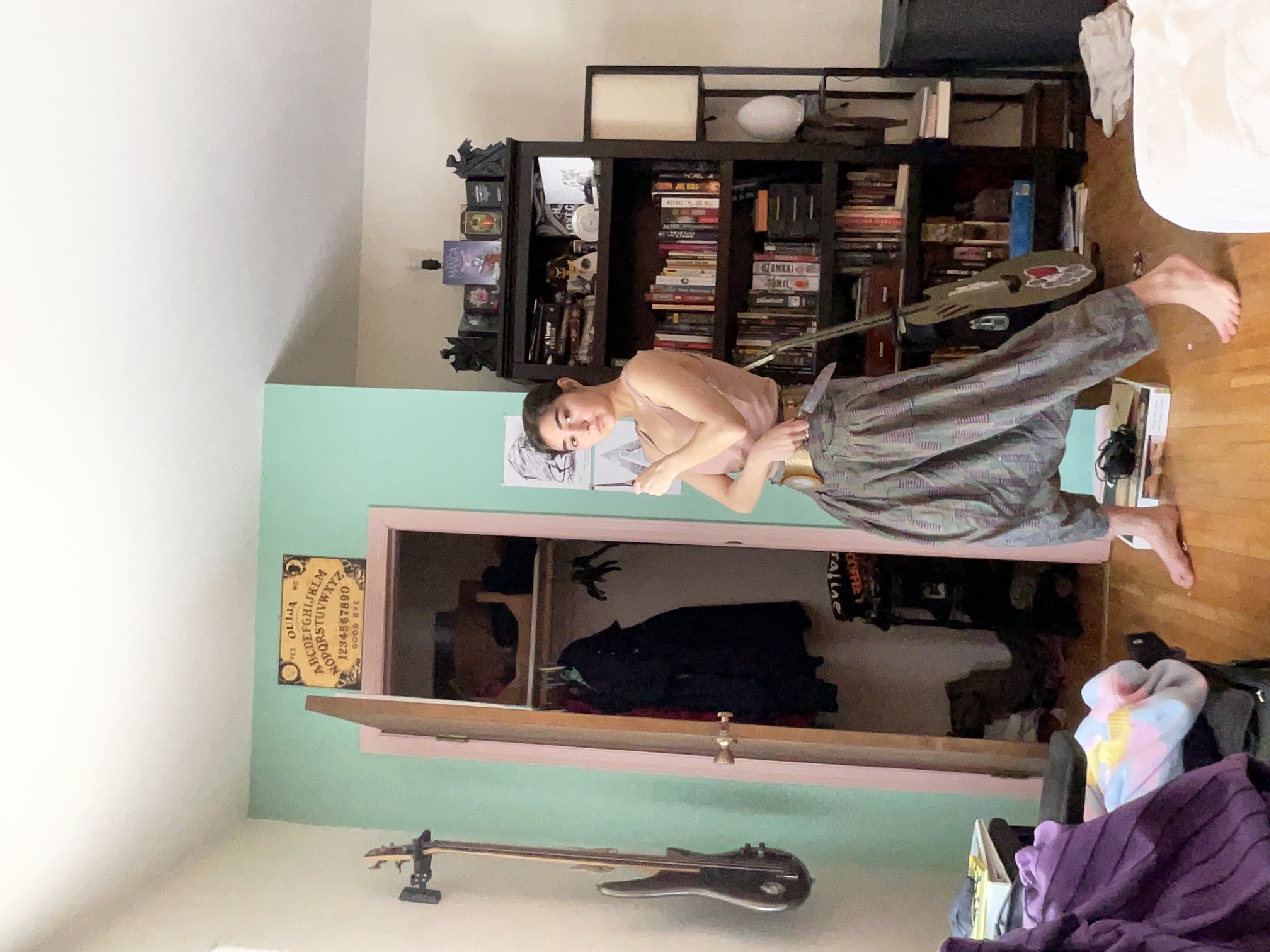a young woman with short brown hair is standing in a bedroom. she's wearing loosefitting, patterned harem pants with a gold belt and a lightcolored sleeveless top.  she's holding what appears to be a guitar or similar instrument. the room has an eclectic and slightly bohemian style, featuring a bookshelf full of books, a closet, a guitar leaning against the wall, and an ouija board decoration on the wall. the overall aesthetic is vintageinspired and somewhat alternative. the room is painted a light teal color. the woman is posed in a relaxed but deliberate manner.
