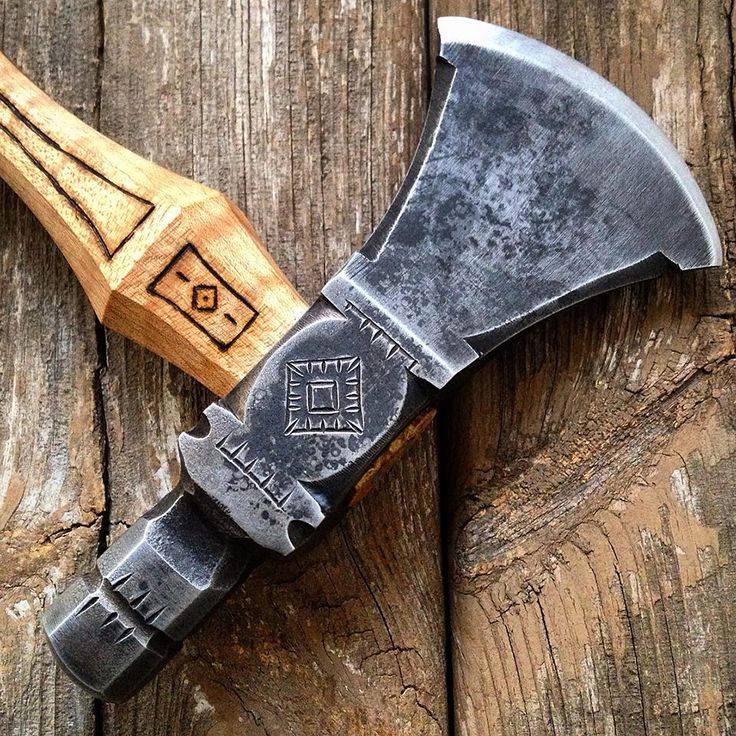 a detailed closeup shows a handforged metal tomahawk lying across a wooden surface. the axe head is weathered and features decorative engravings around its base and along the handle, which has a square emblem etched on it. the axe's handle rests on a weathered wooden plank, and the image focuses on the axe's design and the craftsmanship involved in its creation.