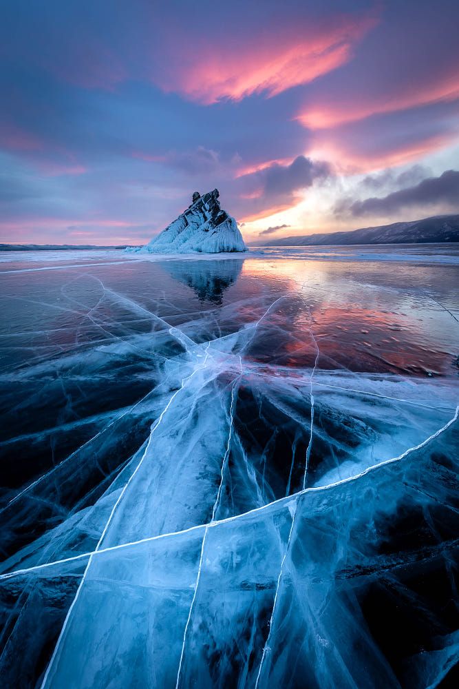 the image captures a scenic view of lake baikal during winter, showcasing a vast expanse of transparent blue ice with prominent cracks across its surface. a rocky island covered in ice stands in the distance, reflecting in the icy surface. the sky is adorned with a colorful display of pink and blue clouds at sunset or sunrise, creating a breathtaking landscape.