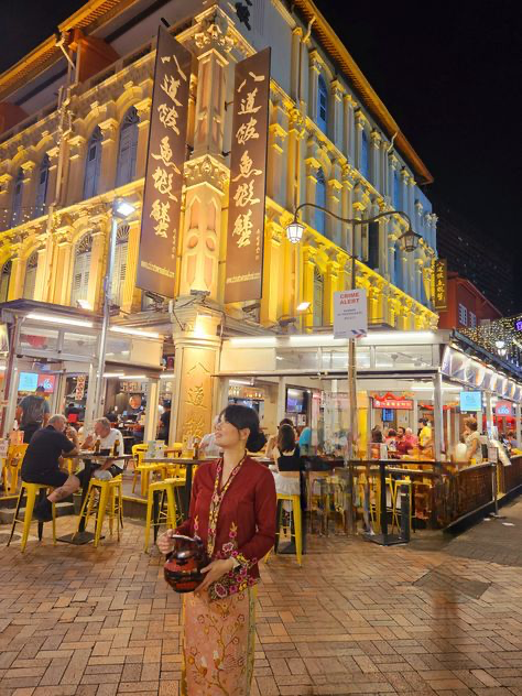 the image shows a vibrant night scene in singapore's chinatown. a woman wearing a traditional baju kurung (malaysian/indonesian dress) stands in front of a brightly lit, ornate building with chinese characters on its facade.  the building houses numerous food stalls and restaurants, with people dining at tables under a covered area. yellow chairs and warm lighting create a bustling atmosphere. the overall scene depicts the rich culture and nightlife of singapore's chinatown.