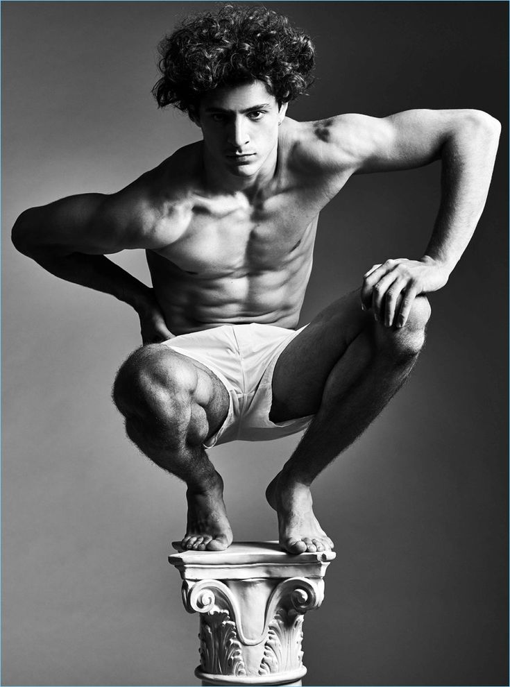 a shirtless, muscular man is sitting on a stone column. he is wearing white shorts. the photo is in black and white. the background is a simple gray studio background.