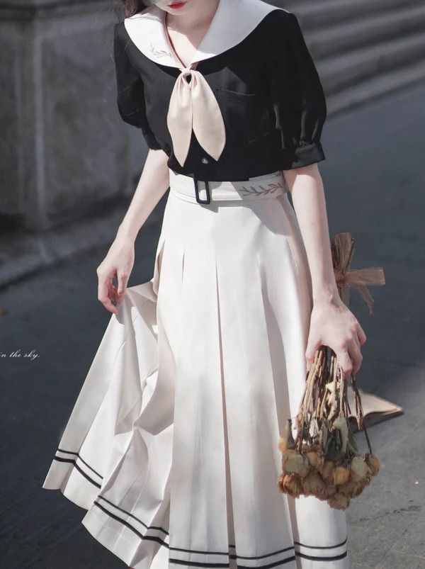 a woman stands on a city street, her back slightly turned towards the camera. she is wearing a vintageinspired outfit with a black shortsleeved top featuring a sailor collar and a white midi skirt. a beige tie and a thin belt cinch her waist. she holds a bouquet of dried flowers in her left hand. the image captures a sense of timeless elegance and urban chic. the bright, natural light illuminates the scene, highlighting the details of her outfit and the texture of the cityscape.