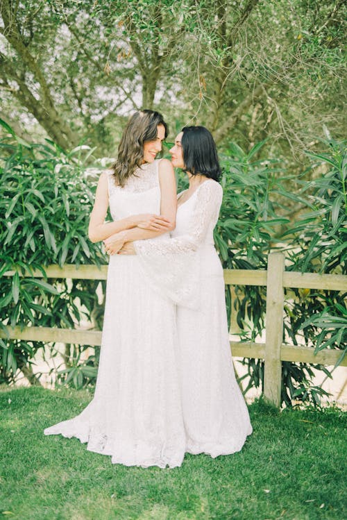 two brides in elegant white lace wedding dresses stand embracing in a lush green garden setting.  they are smiling at each other, showcasing a romantic and happy moment. the background features a wooden fence and abundant greenery, creating a serene and picturesque atmosphere for their special day.  the dresses are long and flowy with delicate lace details. the overall feeling is one of love, joy, and celebration.