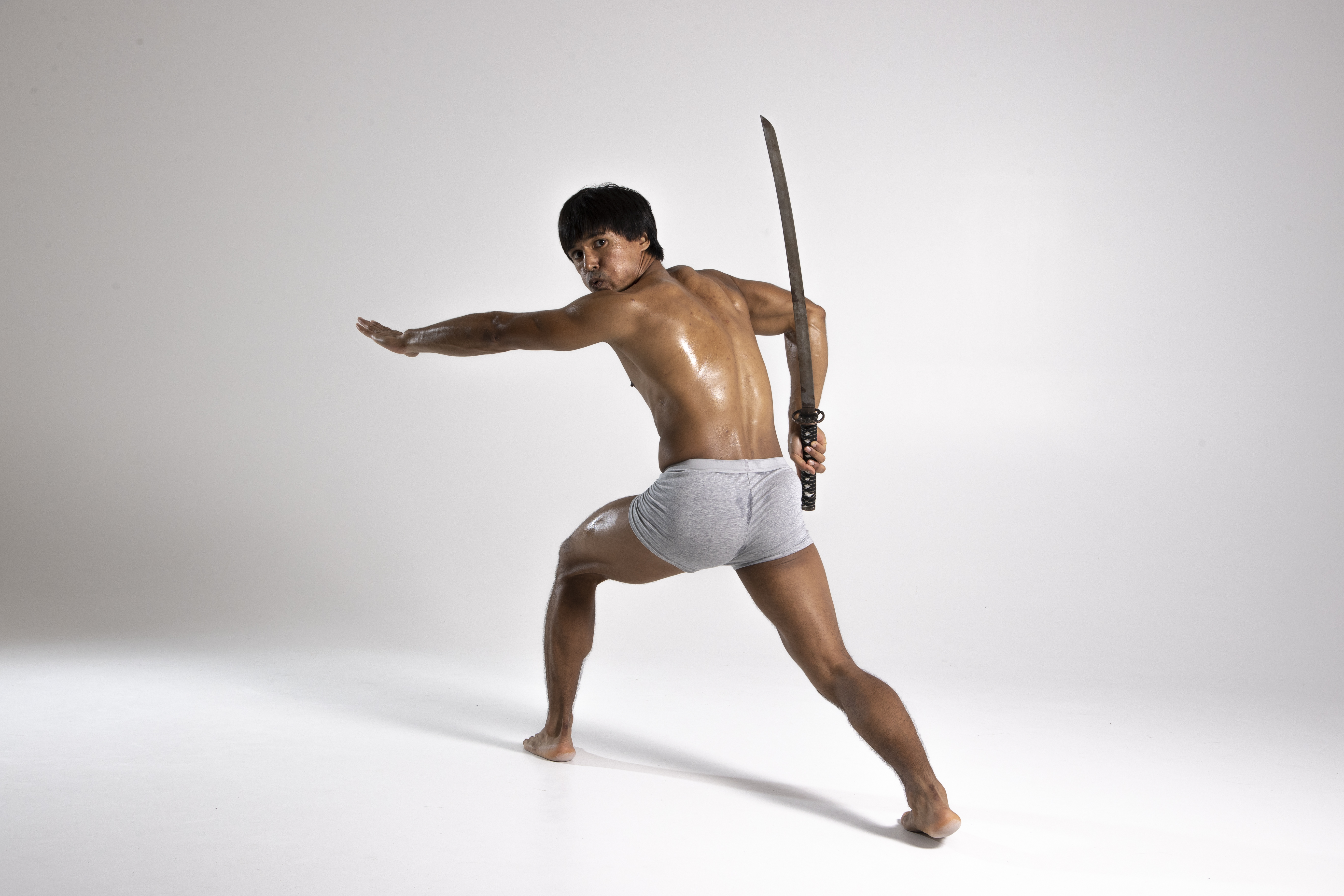 a barechested athletic asian man in gray shorts poses in a studio with a katana sword. his skin appears wet or oiled, and he is in a dynamic stance with one arm extended.