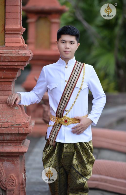 a man dressed in a traditional thai costume. he is standing in front of a temple. he is smiling and looking at the camera. his costume is white with gold embellishments. his sash is also gold and has intricate designs. he is wearing a traditional white longsleeved shirt and gold pants.