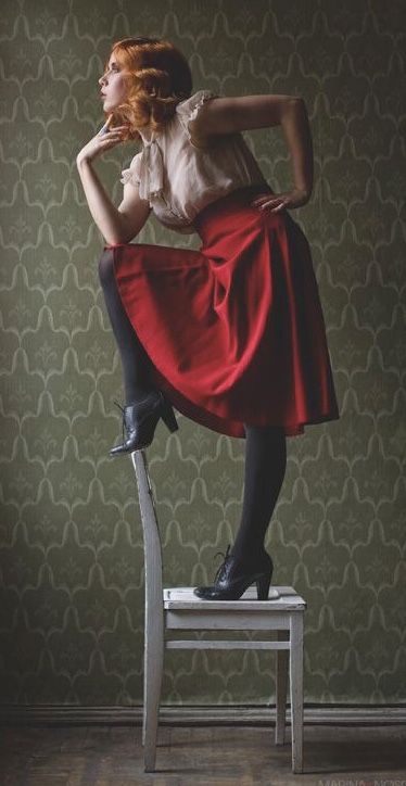 a woman with red hair and a vintage style is posing on a white wooden chair. she's wearing a white blouse, a long red skirt and black shoes. she has her left hand on her hip and the other is near her face. the background is a green patterned wall and the image has a vintage feel to it.