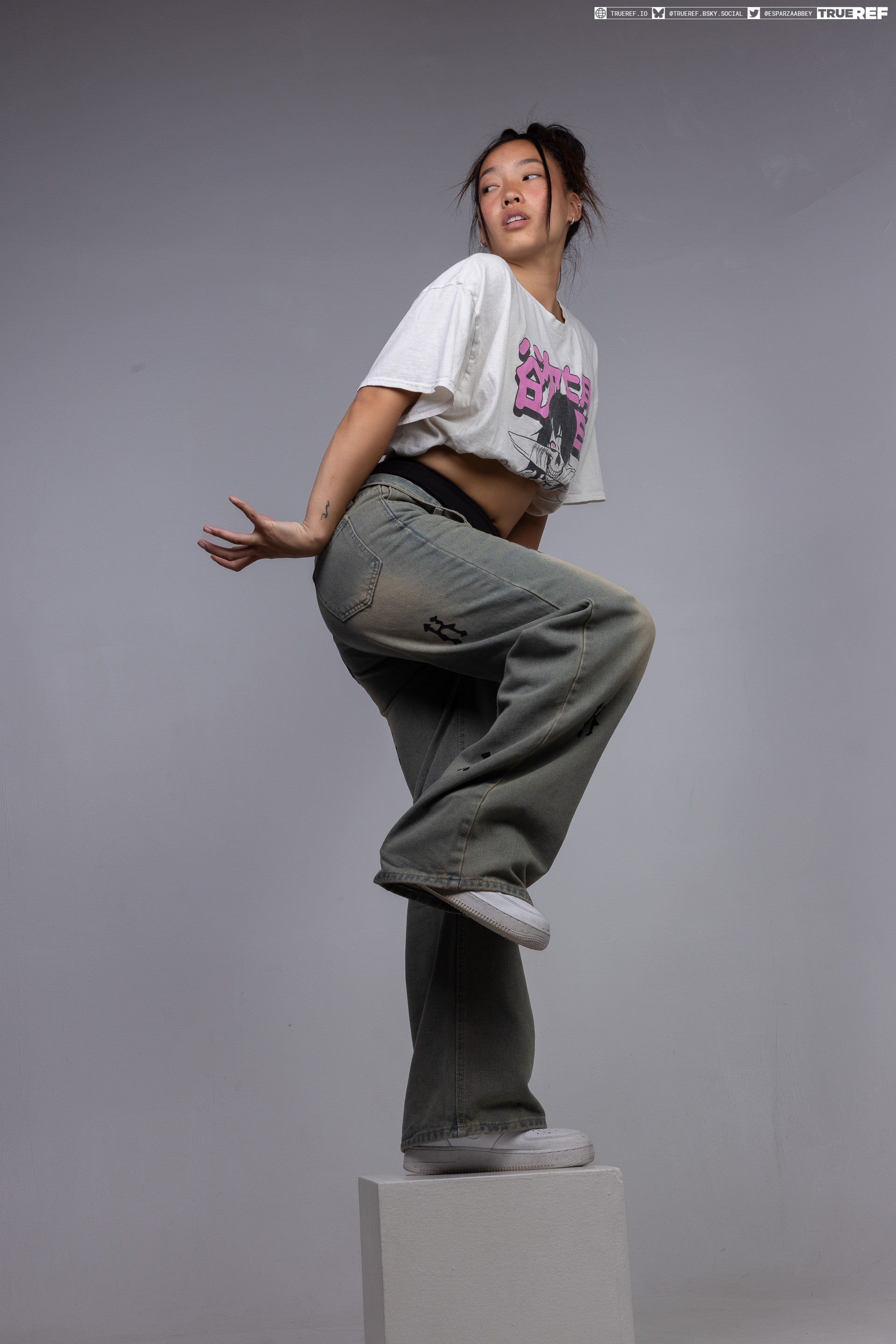 a young asian woman is striking a pose in a studio shot against a gray background. she's standing on a small white platform, her left leg raised with her foot resting on the platform, while her right leg is extended. she's wearing a white tshirt with a graphic on the front, cropped to expose her midriff, and loosefitting, faded jeans. the look is completed with white sneakers. her hair is styled in a messy bun, and she's looking off to the side, away from the camera. the overall impression is casual and fashionable.