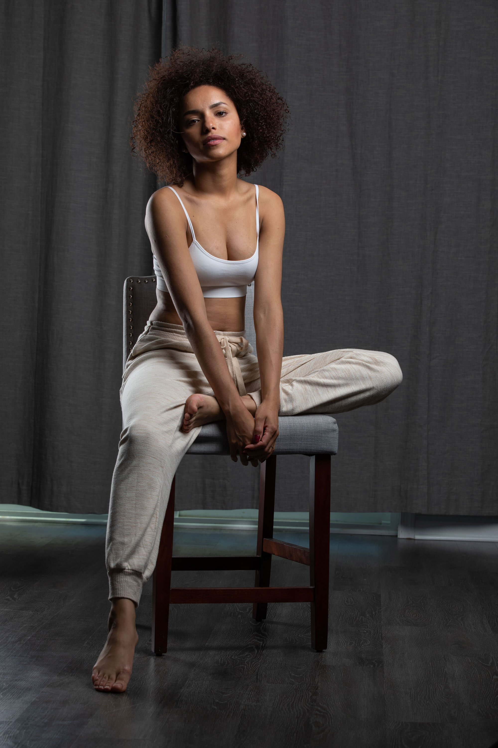 a woman with an afro hairstyle sits on a stool in front of a grey curtain backdrop. she's wearing a white crop top and lightcolored joggers. the lighting on her is soft, and the overall tone of the image is muted.