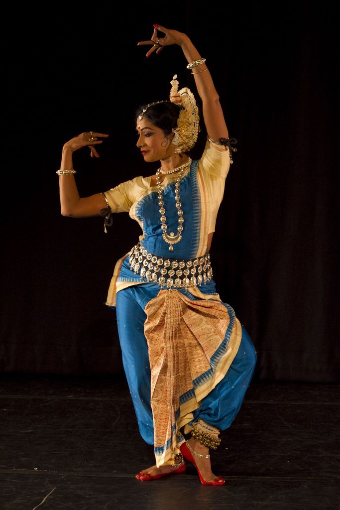 a woman in a traditional indian costume performs a dance on a stage. she is wearing a blue and gold sari with intricate jewelry. she has her arms raised and her hands in a graceful gesture. she is looking to the side and has a serious expression on her face. the stage is dark and the lighting is focused on the dancer.