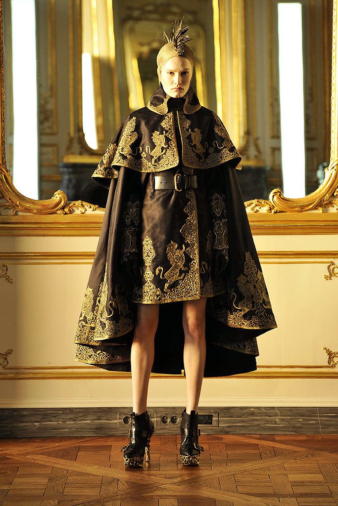 the image showcases a fashion model poised in a striking dark brown gown adorned with elaborate gold embroidery depicting lions. the gown features a dramatic capelike design and is cinched at the waist with a dark brown belt. the model's attire is highly ornate, reminiscent of regal or vintageinspired fashion. she wears high, spiked black boots, completing the look. the backdrop is a richly decorated palace interior with gilded mirrors, creating a sense of grandeur and luxury. the overall aesthetic is high fashion and intensely detailed, likely from a runway show or highend fashion editorial.