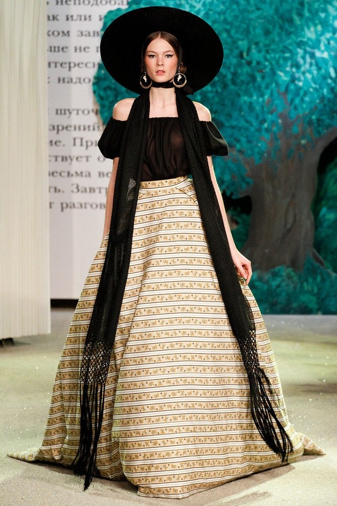 a woman wearing a black and gold striped dress with a black scarf over her shoulders. she has a large black hat with a wide brim, and she is wearing large gold earrings. she is looking down and appears to be walking on a runway. the image is taken from a fashion show.