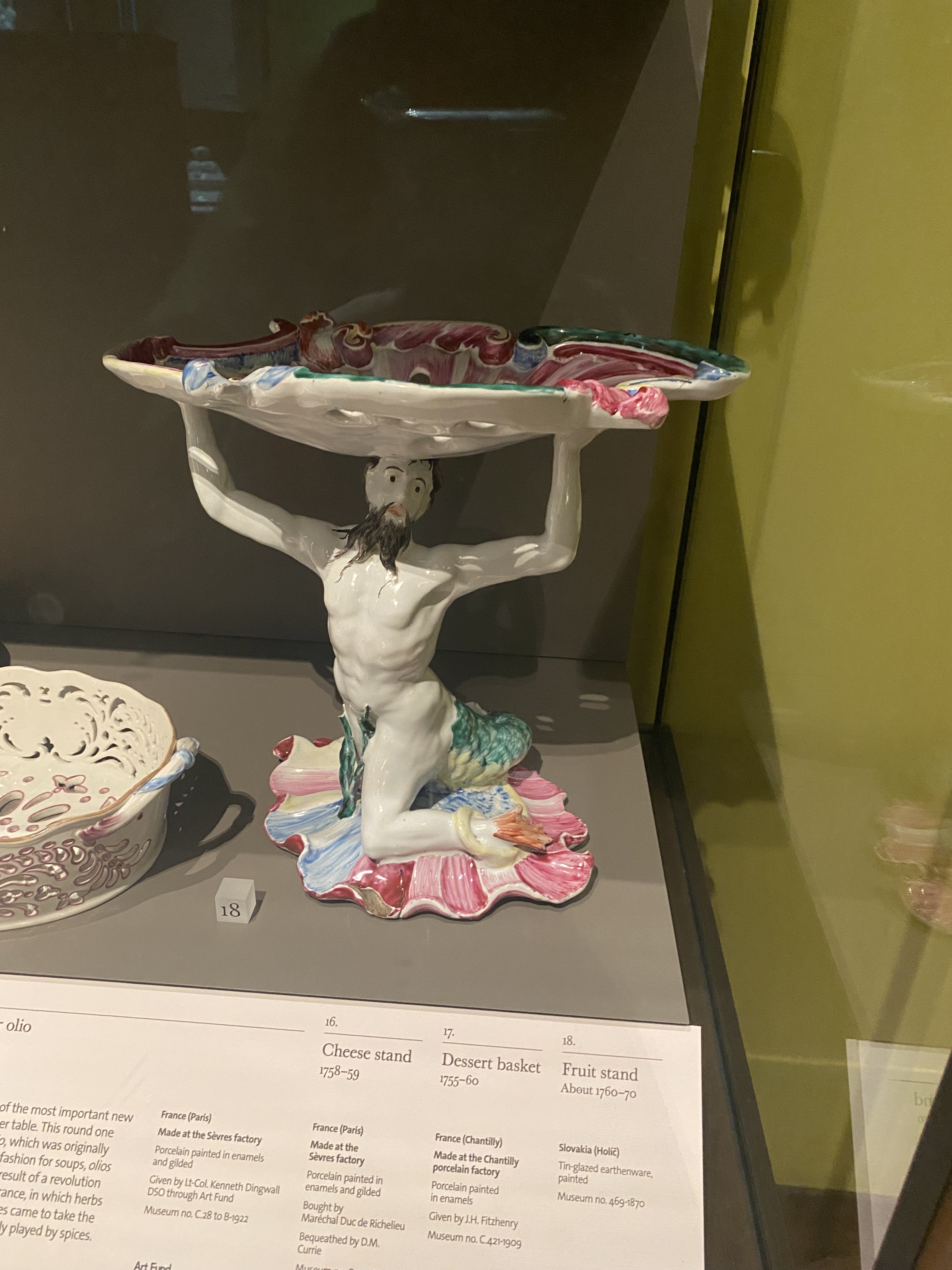 the image shows a porcelain sculpture on display in a museum. the sculpture depicts a man kneeling and holding a large dish above his head. in the background are a display case and descriptive labels for the artifact.