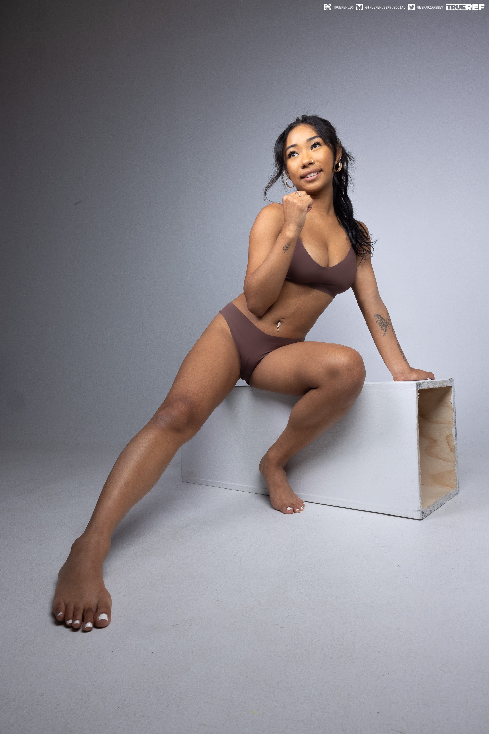 a full shot of an asian woman wearing brown underwear and no shoes. she is squatting on a white box inside a studio. she is barefoot and has fingernail polish on her toes. she has tattoos on her arms. she has hoop earrings and a belly button piercing. she is looking up and posing for the camera.