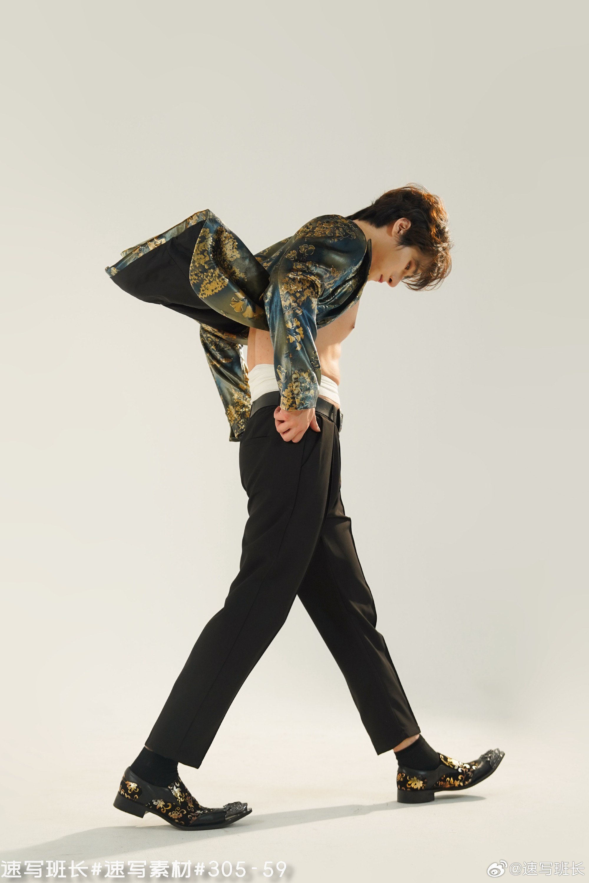 the image is a fashion photo of a young man posing against a white background. the man is wearing black pants, black shoes with floral patterns, and a partially unbuttoned, open jacket with a blue and gold floral pattern. his torso is bare. the lighting is bright and artificial, creating a dramatic and stylish look. the man's pose is dynamic and confident, with his head down and his jacket billowing out behind him.