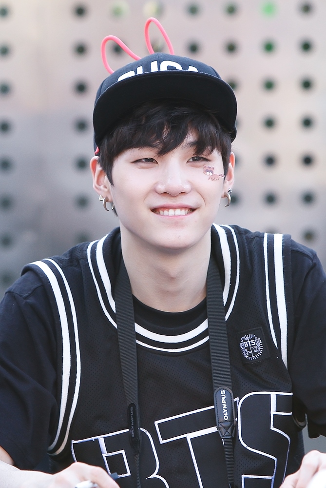 a smiling man, identified as suga from the kpop group bts, is wearing a black baseball cap with pink bunny ears attached. he has dark hair and fair skin. he is also wearing earrings and a black shirt with white stripes, along with a camera strap around his neck. the bts logo is visible on his shirt. he has some decorative markings on his face. the image is taken outdoors.