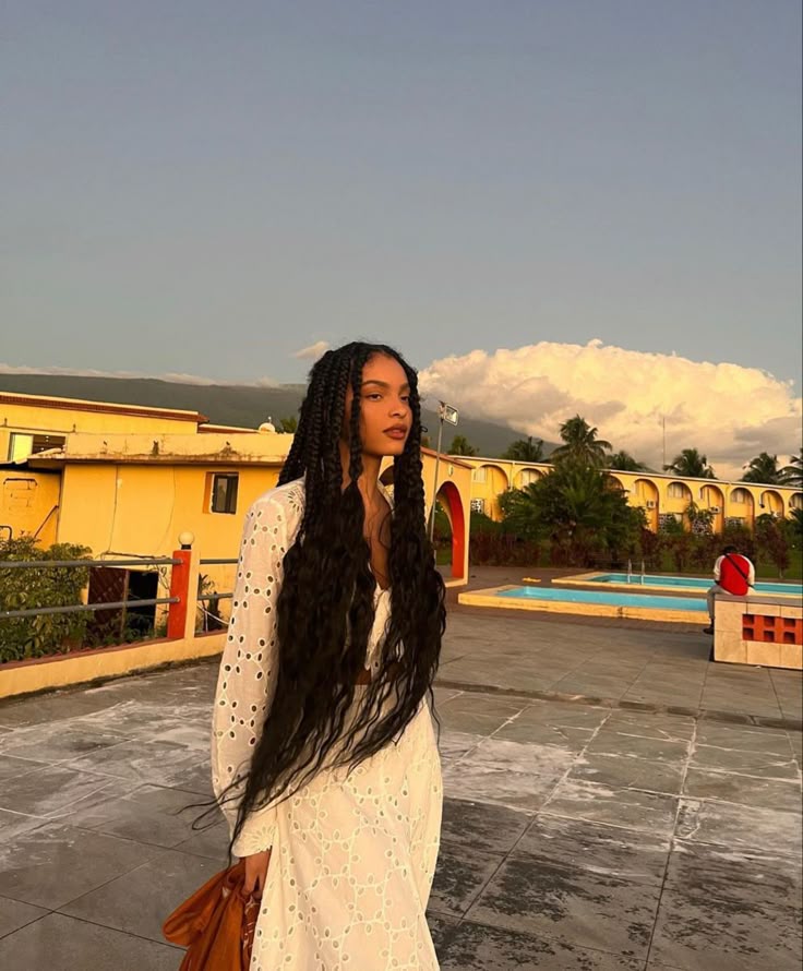 a woman with long braids is standing outdoors in a white dress. behind her is a yellow building, a swimming pool, and a cloudy sky.