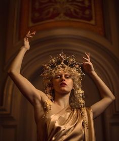 a woman with curly blonde hair is depicted in a golden crown and a draped golden dress. she strikes a pose reminiscent of a classical statue with her arms raised, hands flexed delicately, against a backdrop of an ornate, dimly lit archway. the lighting highlights the gold tones, creating a sense of grandeur and drama.