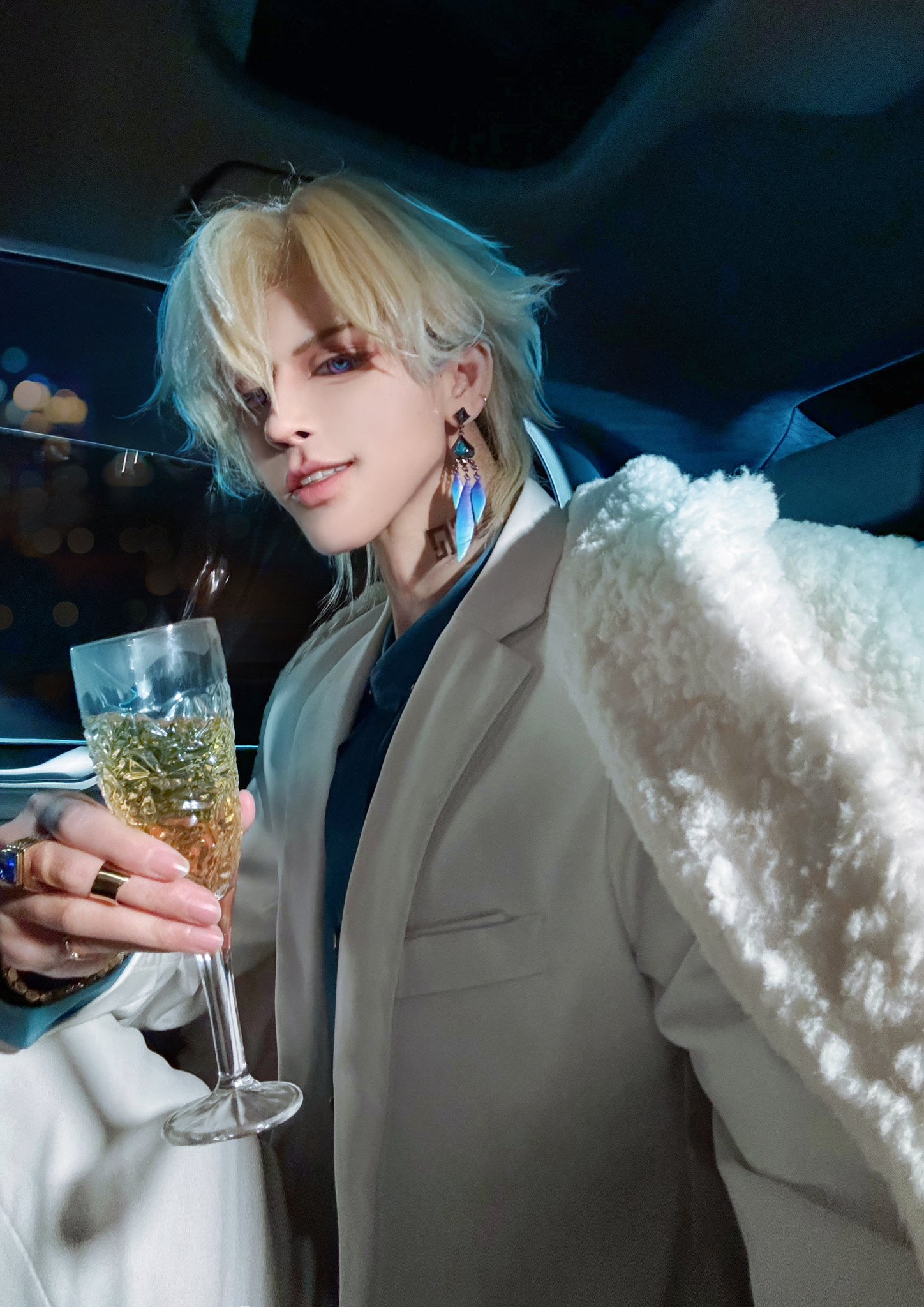 a young man in a white suit with blond hair and blue earrings holds a glass of champagne. he is looking directly at the camera and smiling. he is dressed in cosplay and appears to be in the backseat of a car.