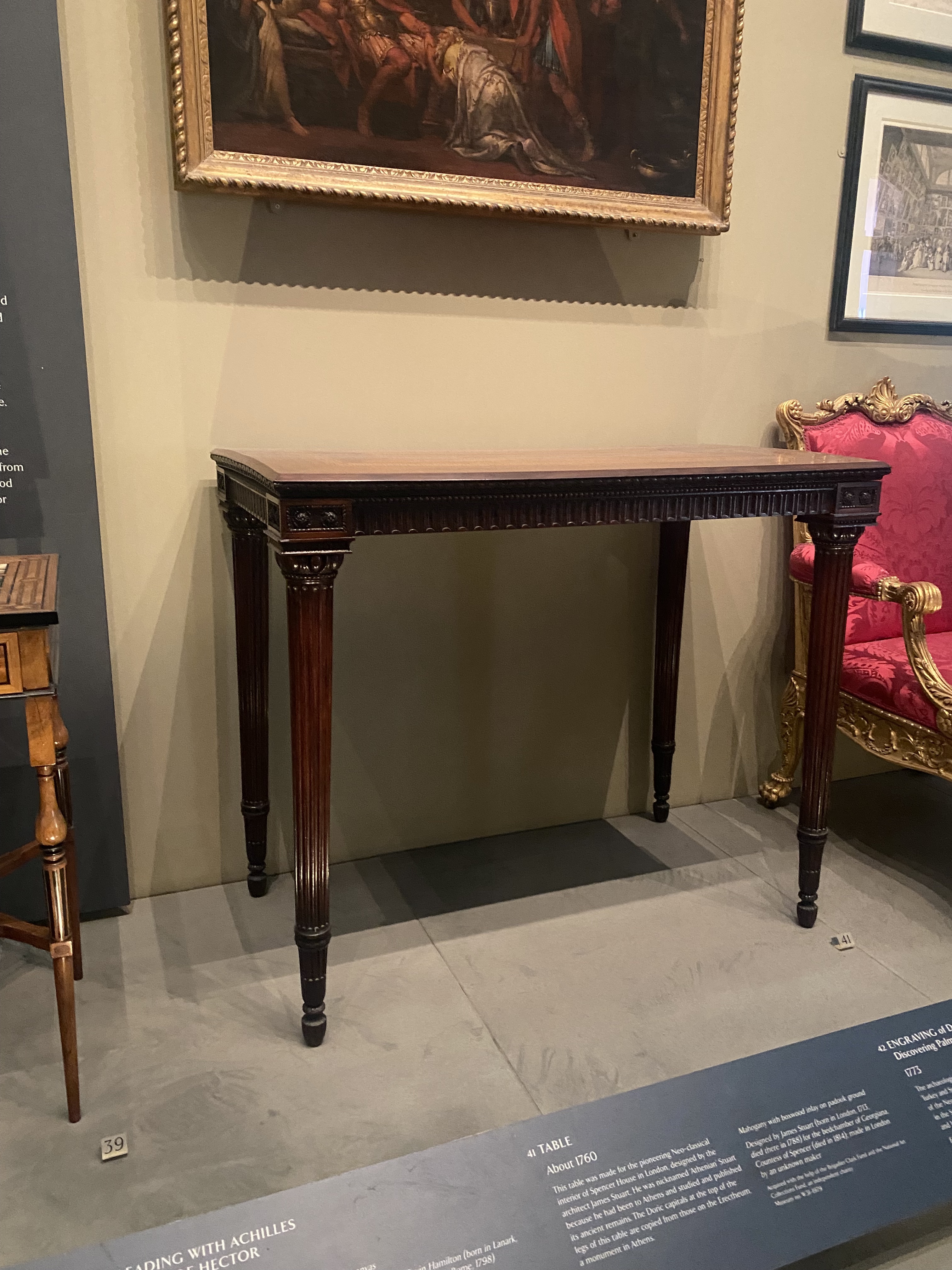 the image shows an antique mahogany table on display in a museum. a painting hangs above it on the wall. there is another piece of furniture, possibly a chair, partially visible to the right, and another piece of furniture to the left of the table. a museum display sign is partially visible in the foreground.