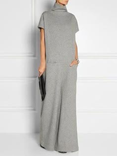 a woman is standing in a grey dress. the dress has a turtleneck, short sleeves, and is loose fitting. she is holding a black clutch in her hand.
