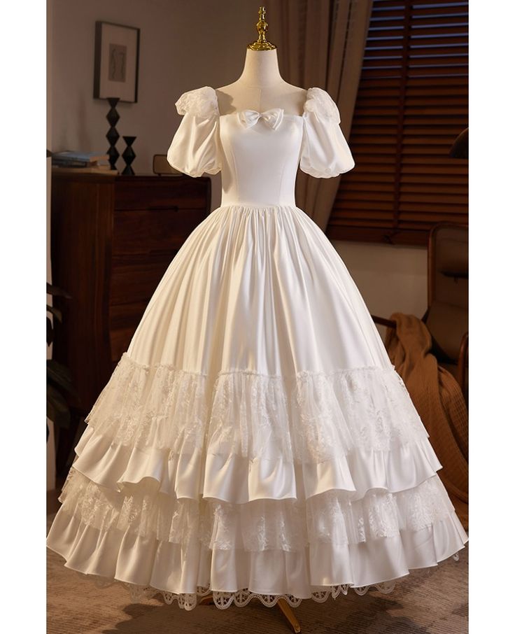 a fulllength image showcasing an elegant, vintageinspired white wedding gown displayed on a mannequin. the dress features a sweetheart neckline, puffed short sleeves, and a full, layered skirt adorned with intricate lace detailing. the backdrop reveals a glimpse of a welllit room with a wooden floor, a chest of drawers, and window blinds.
