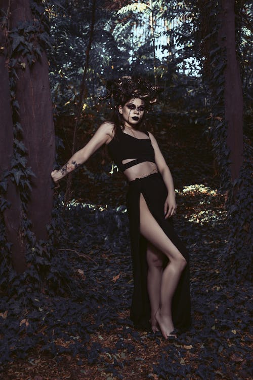 the image showcases a woman dressed in a dark gothic fantasy costume. she wears a black crop top and a long black skirt with a high slit, revealing one leg.  a dark horn headpiece adorns her head, and her makeup is dramatic and dark.  she stands in a dark, shadowy forest setting, leaning against a tree. the overall mood is mysterious and evocative of a witch or sorceress. the lighting and the woman's pose are carefully composed for a dramatic effect.