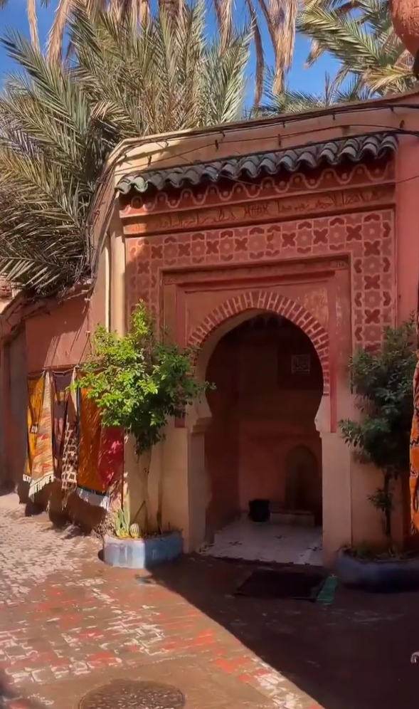 this image shows a view of an ornately decorated courtyard in what appears to be a traditional moroccan or north african building, likely in marrakech. the architecture features a salmoncolored arched doorway with intricate geometric tilework above it. the doorway leads into a recessed area with a lightcolored floor, and a dark object can be seen further inside. the building's exterior is surrounded by lush plants, including small trees in planters and tall palm trees in the background, which adds to the overall exotic atmosphere. on the left side of the frame, colorful rugs are hung, contributing to the vibrant and cultural feel of the scene. the courtyard is paved with a redbrick pattern, and the image captures the warm, sunny lighting typical of the region.