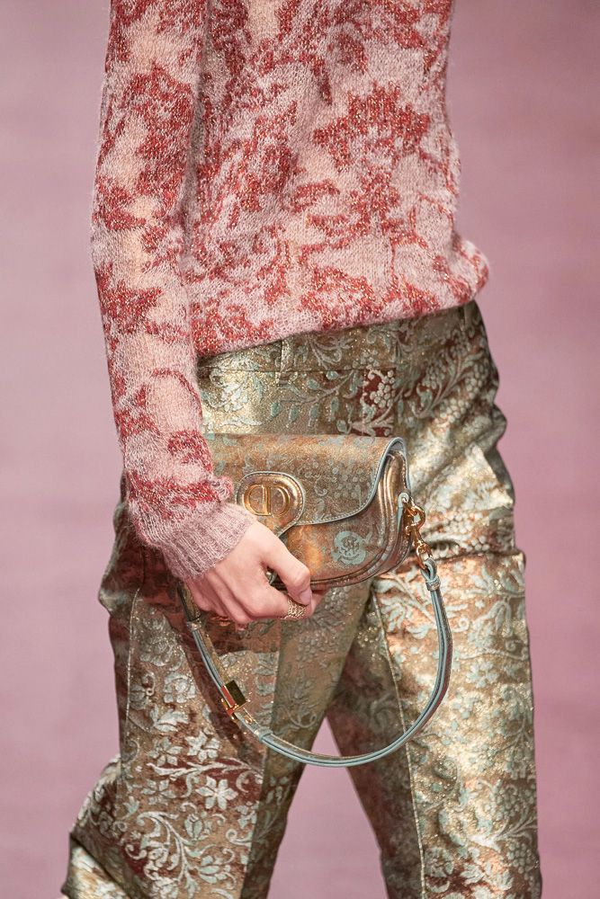 closeup shot of a woman's outfit featuring a red and beige patterned knit sweater and gold brocade pants. she's carrying a metallic gold and teal handbag with a light teal strap. the focus is on the textures and patterns of the clothing and the bag. the background is blurred out, indicating a runway or fashion shoot setting. the overall style is autumnal or winter, with rich textures and luxurious materials.
