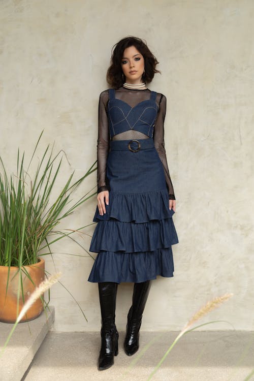 a woman stands in front of a tan wall wearing a denim twopiece outfit, consisting of a crop top and a tiered skirt with a belt. she also wears a black mesh top underneath and black patent leather kneehigh boots. a string of pearls adorns her neck. to her left, there is a potted plant.