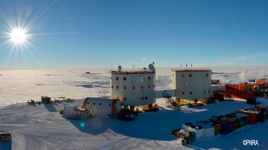 a wideangle shot of a research station in antarctica, showcasing a snowy landscape and a bright sun in the sky. two cylindrical buildings are the focal point, with numerous other structures scattered around them. the station is surrounded by a vast expanse of white snow and ice, highlighting the extreme conditions of the antarctic environment. the image captures a sense of isolation and the important work being conducted in this remote location.